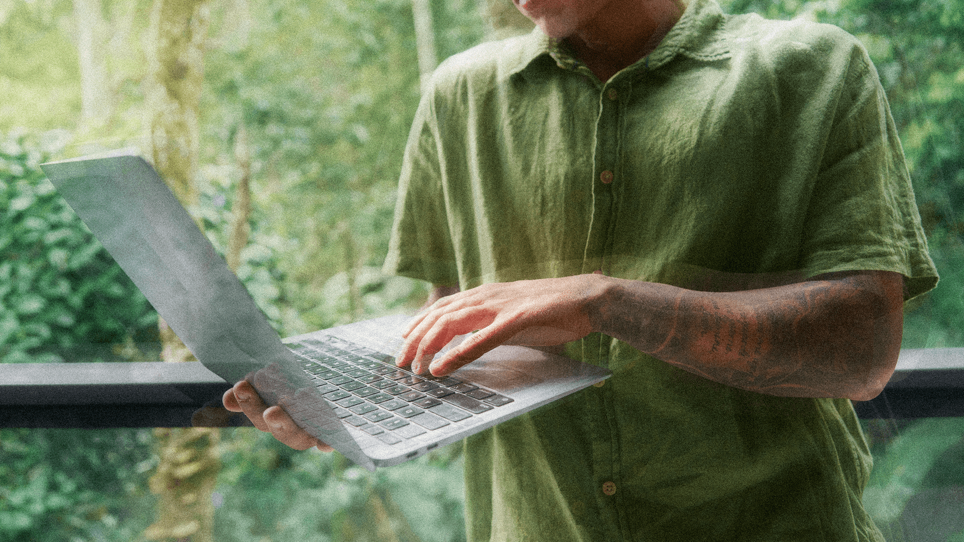 A person wearing a green shirt stands and types on a laptop in front of a lush, green forest backdrop.