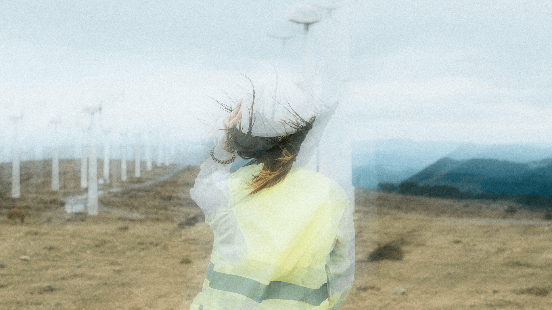 Woman in a safety vest and hard hat facing wind turbines on a hillside