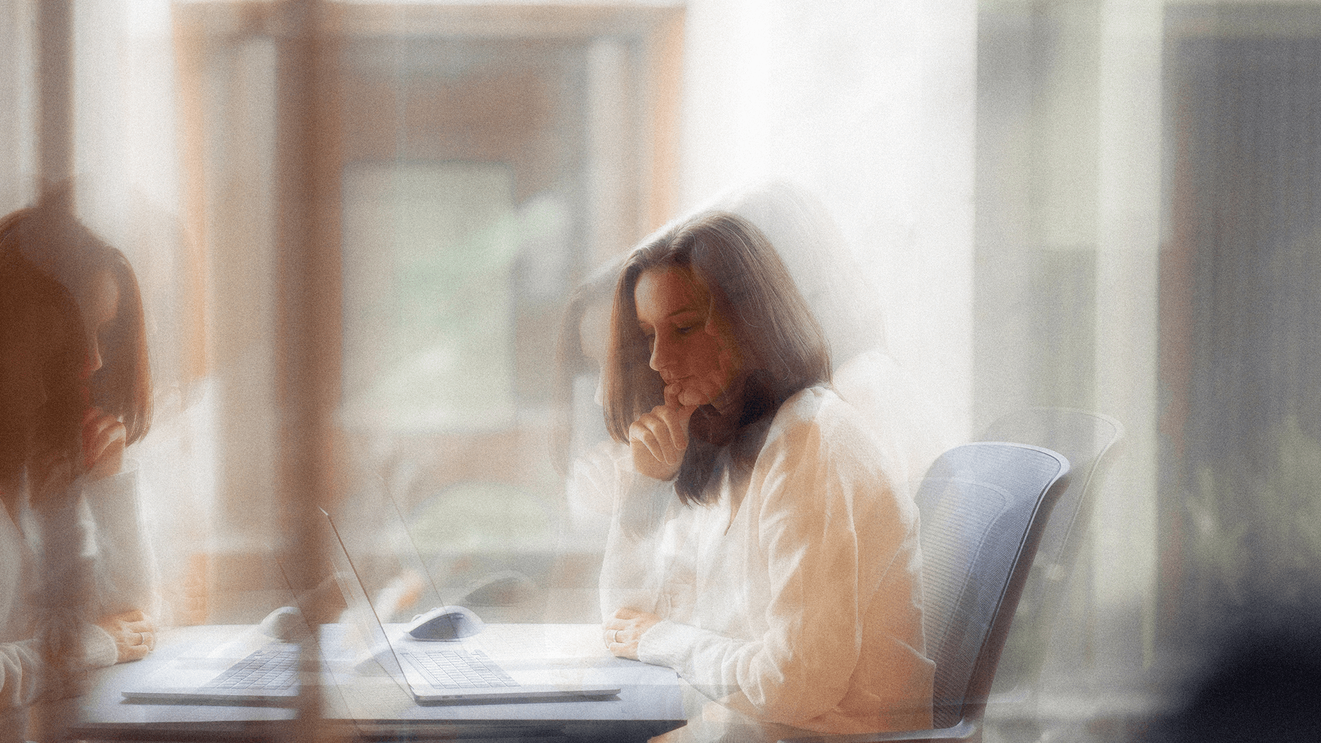 A woman sitting at a desk with a laptop, resting her chin on her hand and looking at the screen, captured with a layered motion effect.