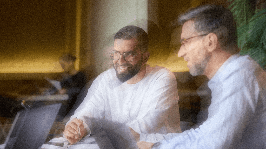 Two men in conversation at a table with documents and laptops