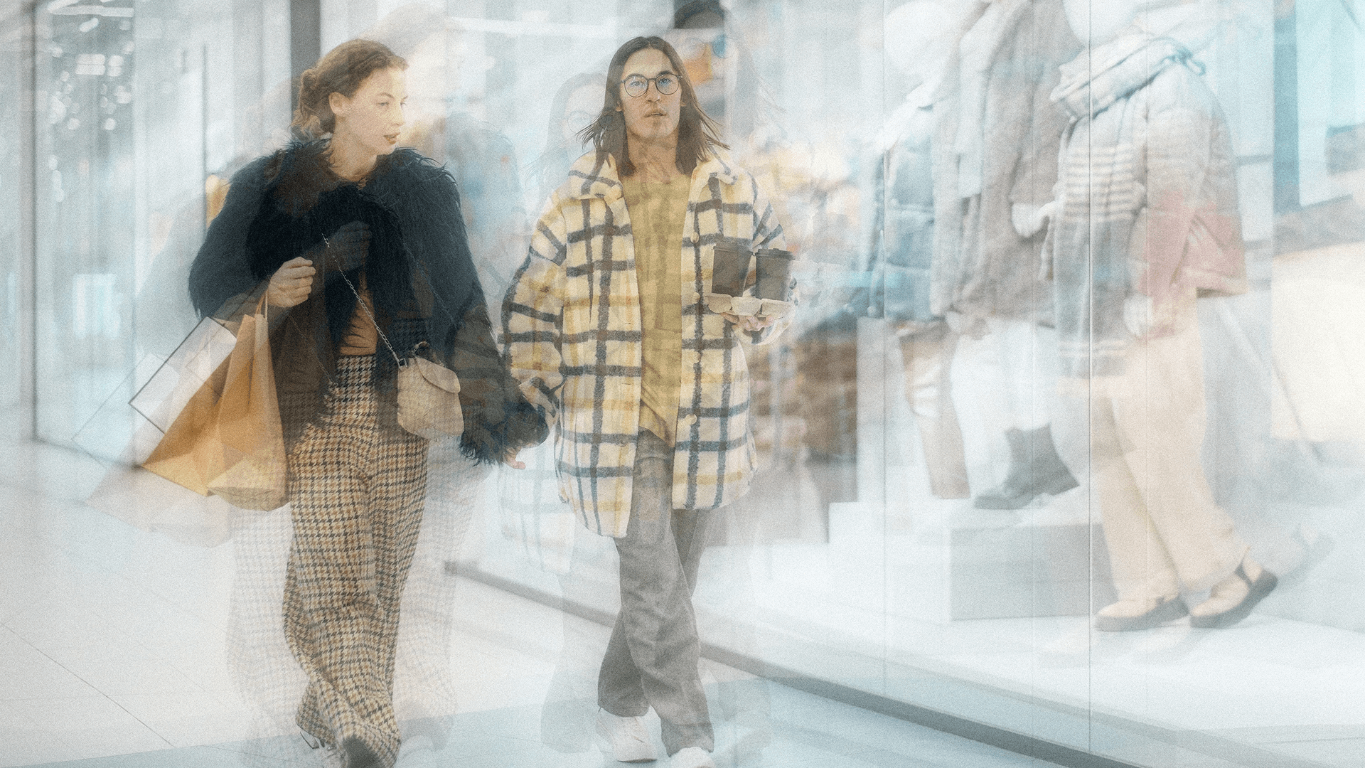 Two people holding hands walking through a shopping mall, holding drinks and bags, with shop windows and mannequins in the background.