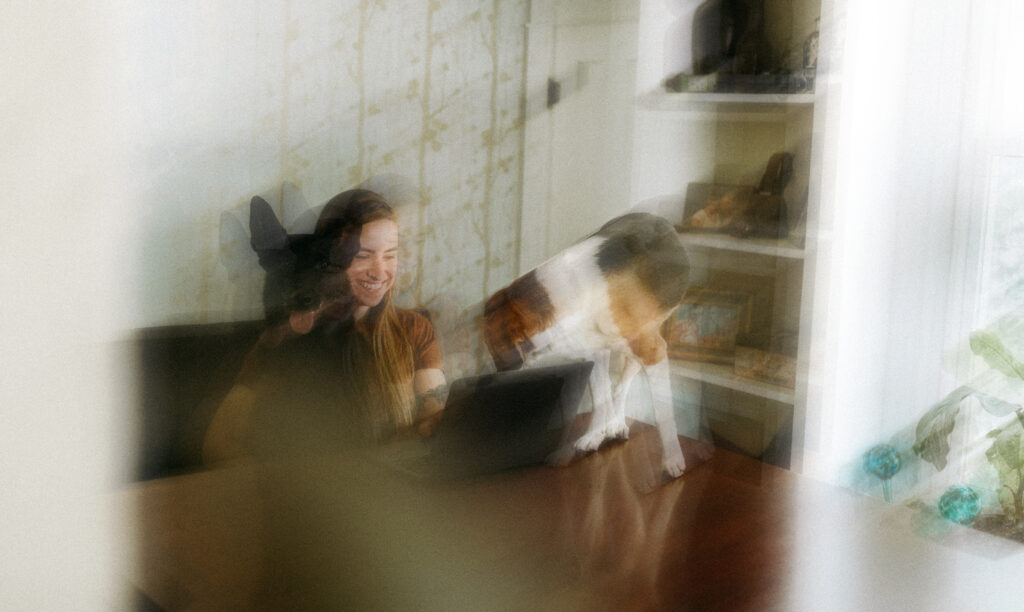 Woman smiling while working on a laptop at home with two dogs nearby, in a dreamy, motion-blurred setting.